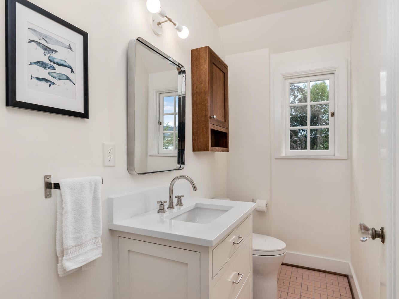 A small bathroom in Portland, Oregon, with white walls features a framed picture of fish, a rectangular mirror, and a sink with a silver faucet. A wooden cabinet and towel hang on the wall next to a window above the toilet offering views of lush greenery.