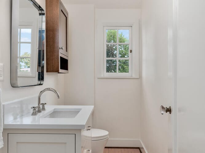 A narrow bathroom in Portland, Oregon, features a white vanity with a rectangular mirror and a modern light fixture above. A wall-mounted cabinet sits nearby, while the toilet is next to a small window framing an outdoor view. The tiled floor is accented by a patterned rug.