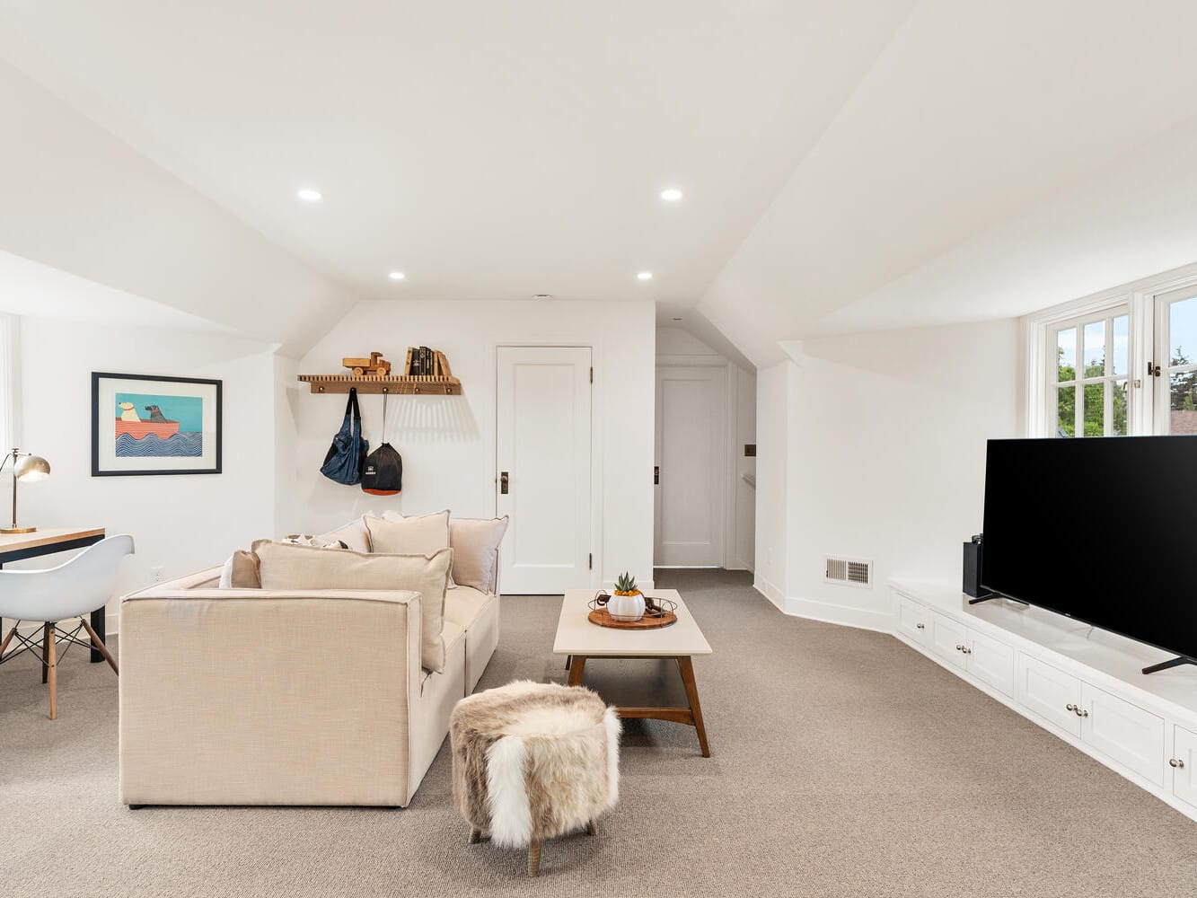 A cozy, modern attic living room in Portland, Oregon, features beige carpet and white walls. It boasts a sofa, coffee table, TV on a low cabinet, a desk with a chair by the window, and a small shelf with hooks on the wall. A colorful framed picture adds a pop of color.