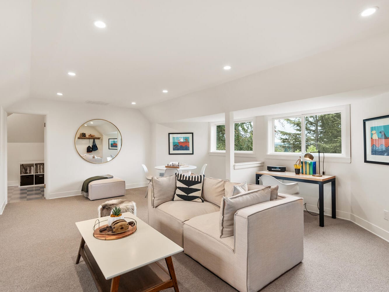 A bright, modern living room in Portland, Oregon, boasts a beige sofa, white coffee table, and wall art. The space features a round mirror, a desk near large windows, and overhead recessed lighting, with a cozy seating area in the corner.