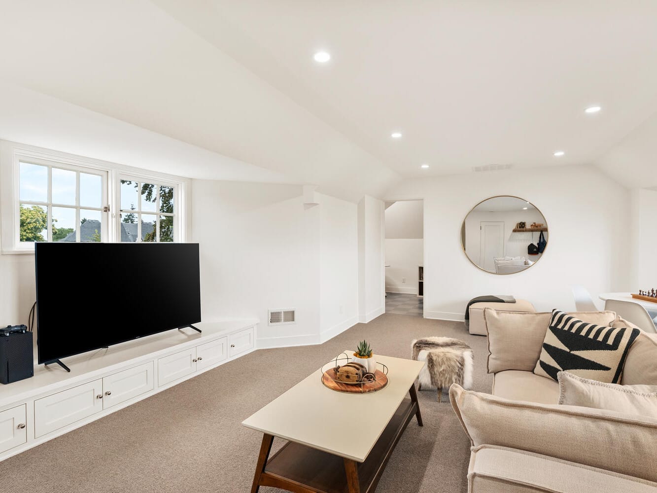 A spacious living room with white walls and carpet, typical of Portland, Oregon homes, featuring a large flat-screen TV on a cabinet, a beige sofa with patterned cushions, a wooden coffee table, and a round mirror on the wall. There's indirect lighting and a glimpse of the dining area.