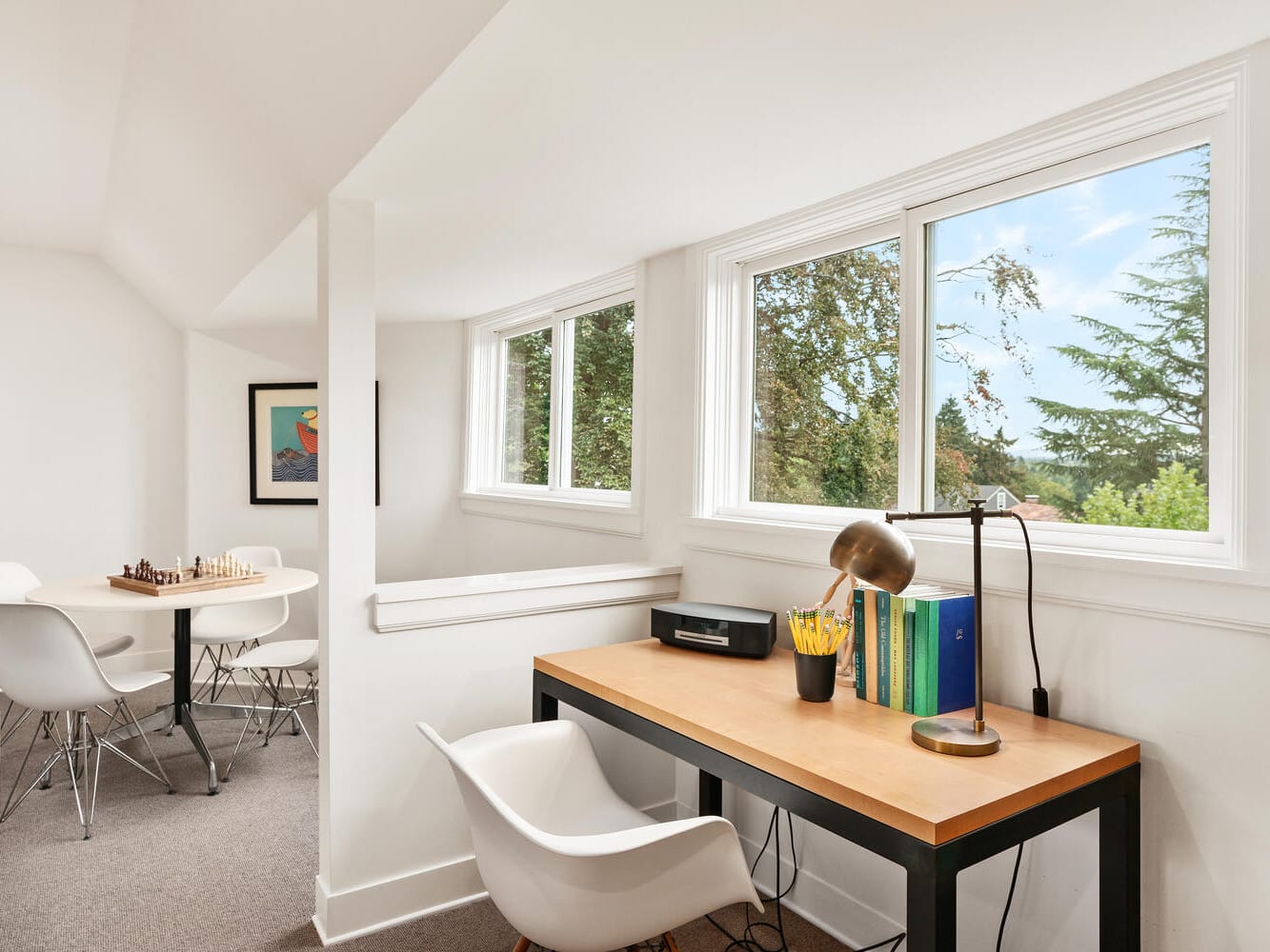 Bright home office with a wooden desk, white chair, and a lamp. Books and a printer sit on the desk. Large windows overlook the lush trees of Portland, Oregon. In the background, a round table with white chairs and a chessboard under a modern art print enhances the space.