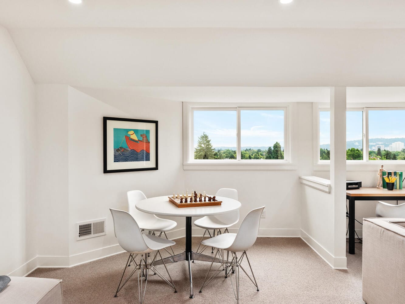 A bright room in Portland, Oregon features a round table with four white chairs and a chess set. A vibrant framed art piece hangs on the wall. Large windows offer a scenic view, and a desk to the right holds a lamp and books, blending perfectly with the neutral decor for a calm atmosphere.