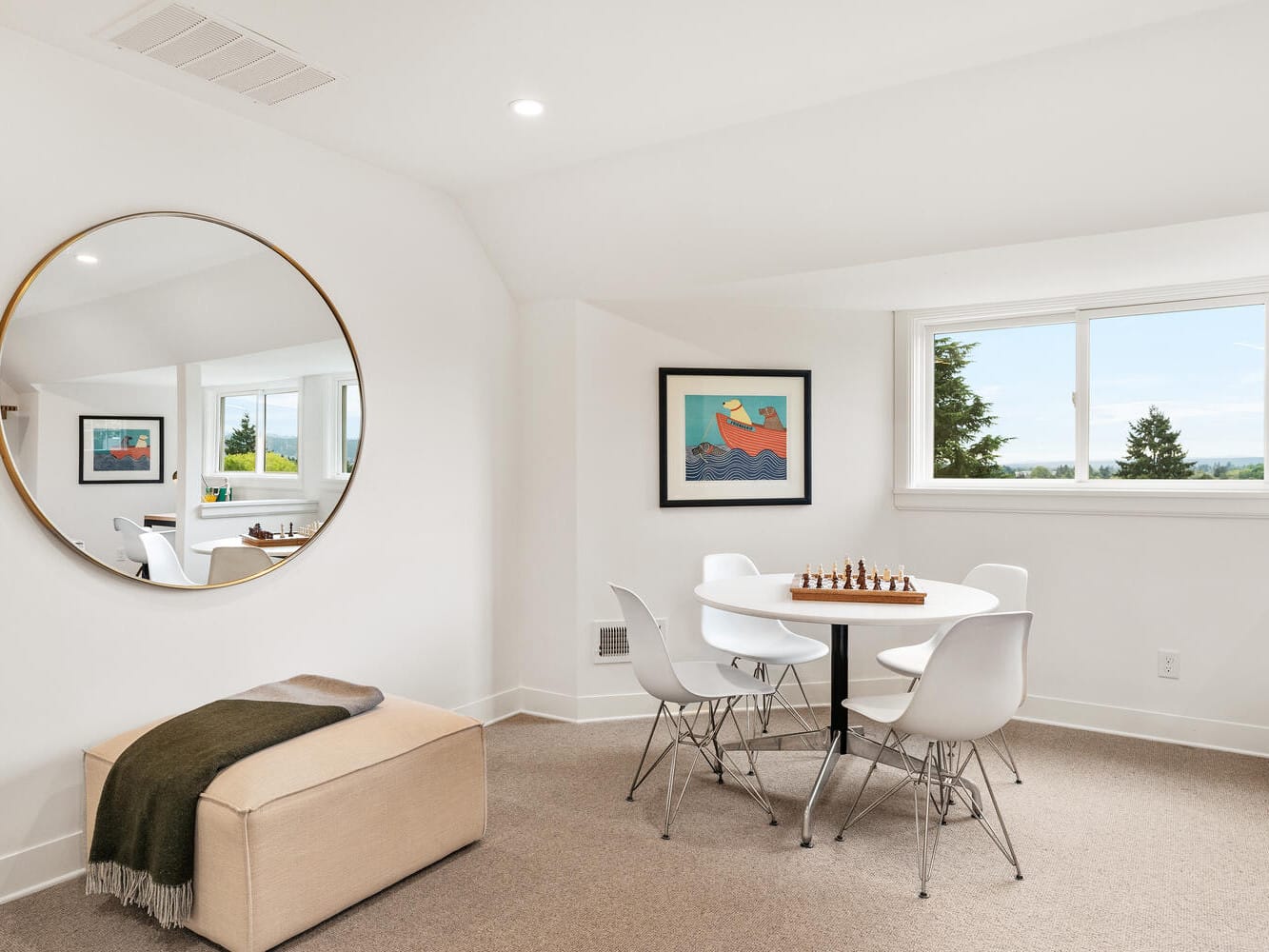 A cozy room in Portland, Oregon features a round mirror reflecting a table with four white chairs and a chess set. A framed artwork adorns the wall above, while a beige ottoman with a green throw sits nearby, completing this inviting space with its large window offering a scenic view.