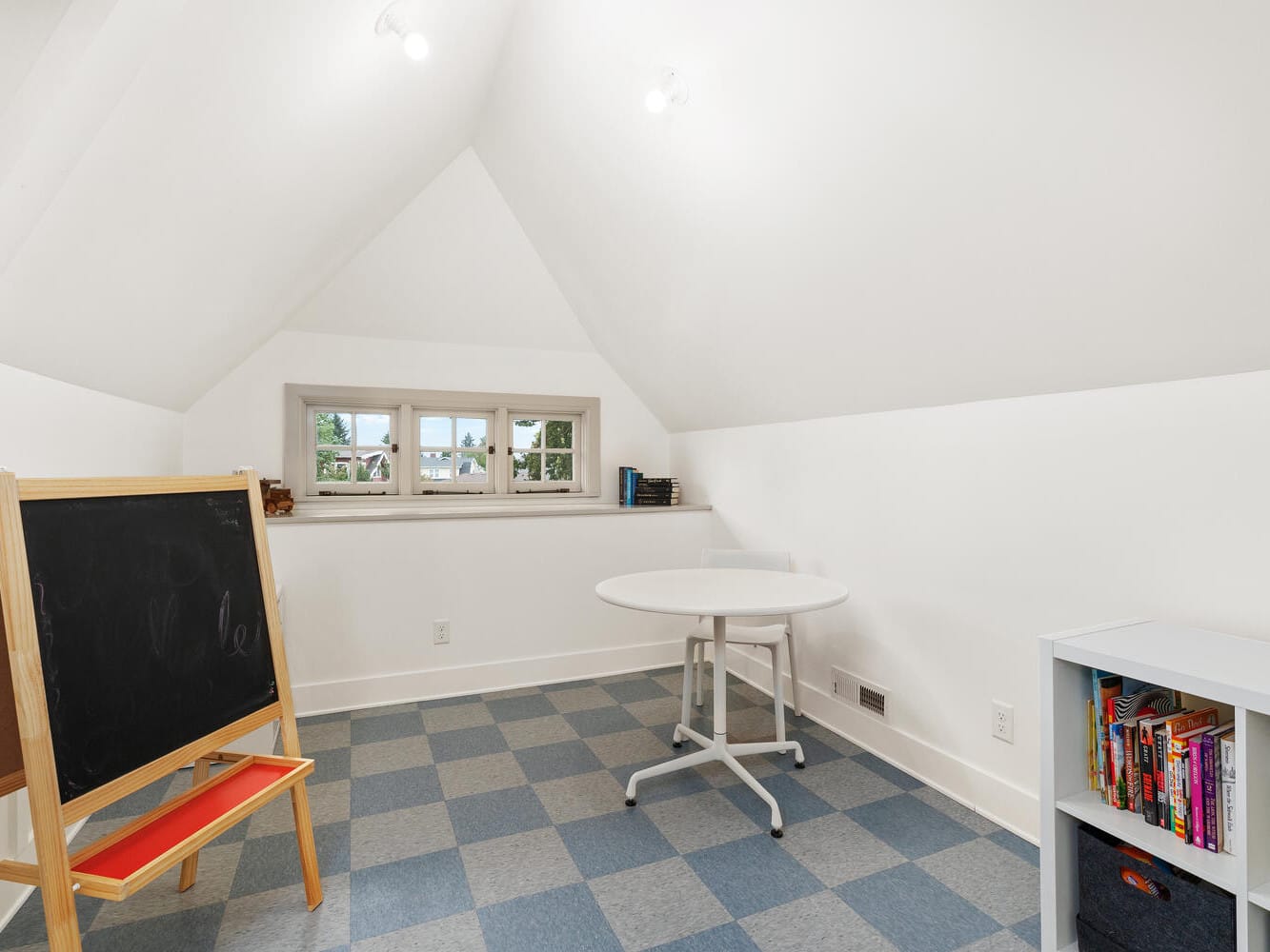 A bright attic space in Portland, Oregon, with a checkered blue and white floor features a small round table, a bookshelf with assorted books, and a blackboard on an easel. The room has white walls and a small window letting in natural light.