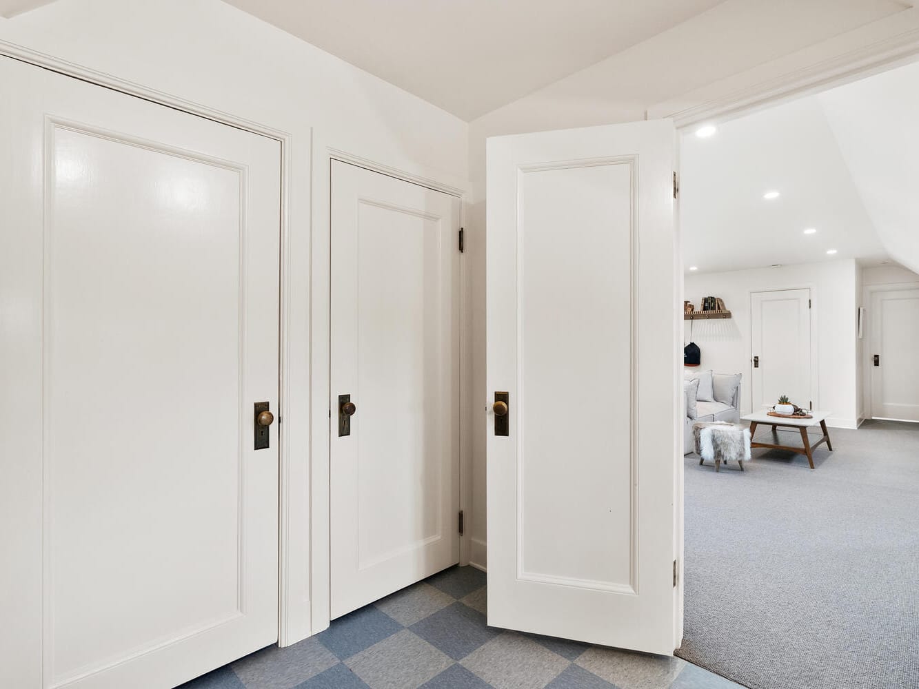 A room in Portland, Oregon, features white walls and three open doors. Beyond, a gray carpet hosts a small table with decorative items, a shelf, and a light-colored armchair. The foreground boasts an eye-catching checkered floor pattern.