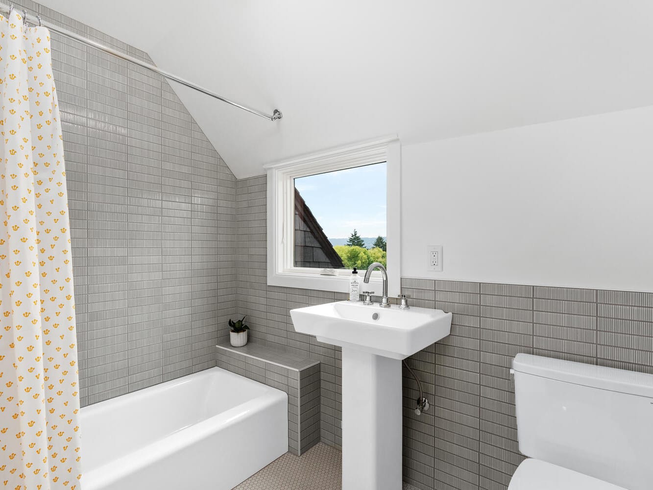 A bright, modern bathroom in a Portland, Oregon home features gray tiled walls, a white bathtub with a yellow-patterned shower curtain, a pedestal sink, and a small plant on the ledge. A window offers an inviting view of trees and the clear sky.