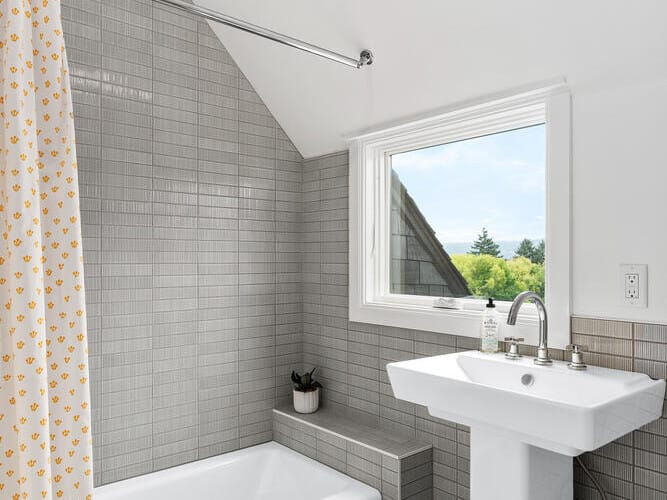 A modern bathroom in Portland, Oregon, boasts gray tiles and features a white bathtub with a yellow patterned shower curtain. Complemented by a white pedestal sink and a large window overlooking lush greenery, the space feels bright and airy beneath its sloped ceiling.