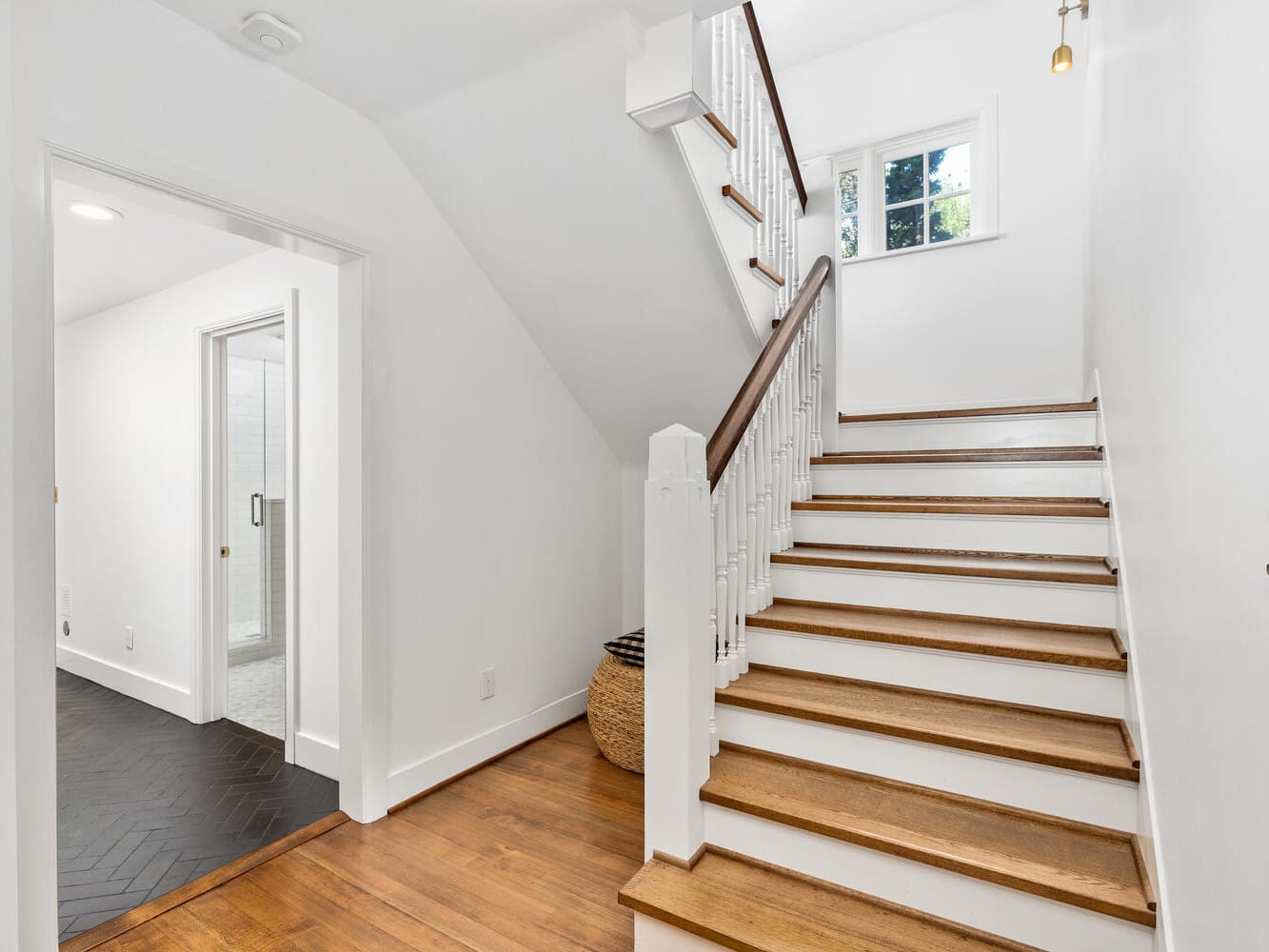 A bright hallway in Portland, Oregon, features a wooden staircase with white railings leading to an upper floor. Natural light streams through a window, and an open door reveals a bathroom with dark herringbone-patterned flooring.