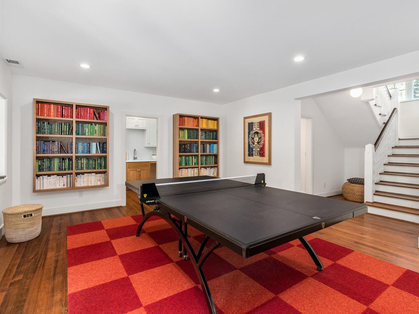 A bright game room in Portland, Oregon features a ping pong table on a red checkered rug. Shelves filled with colorful books line the walls. A doorway leads to a kitchenette, and stairs with a basket underneath ascend to the right.