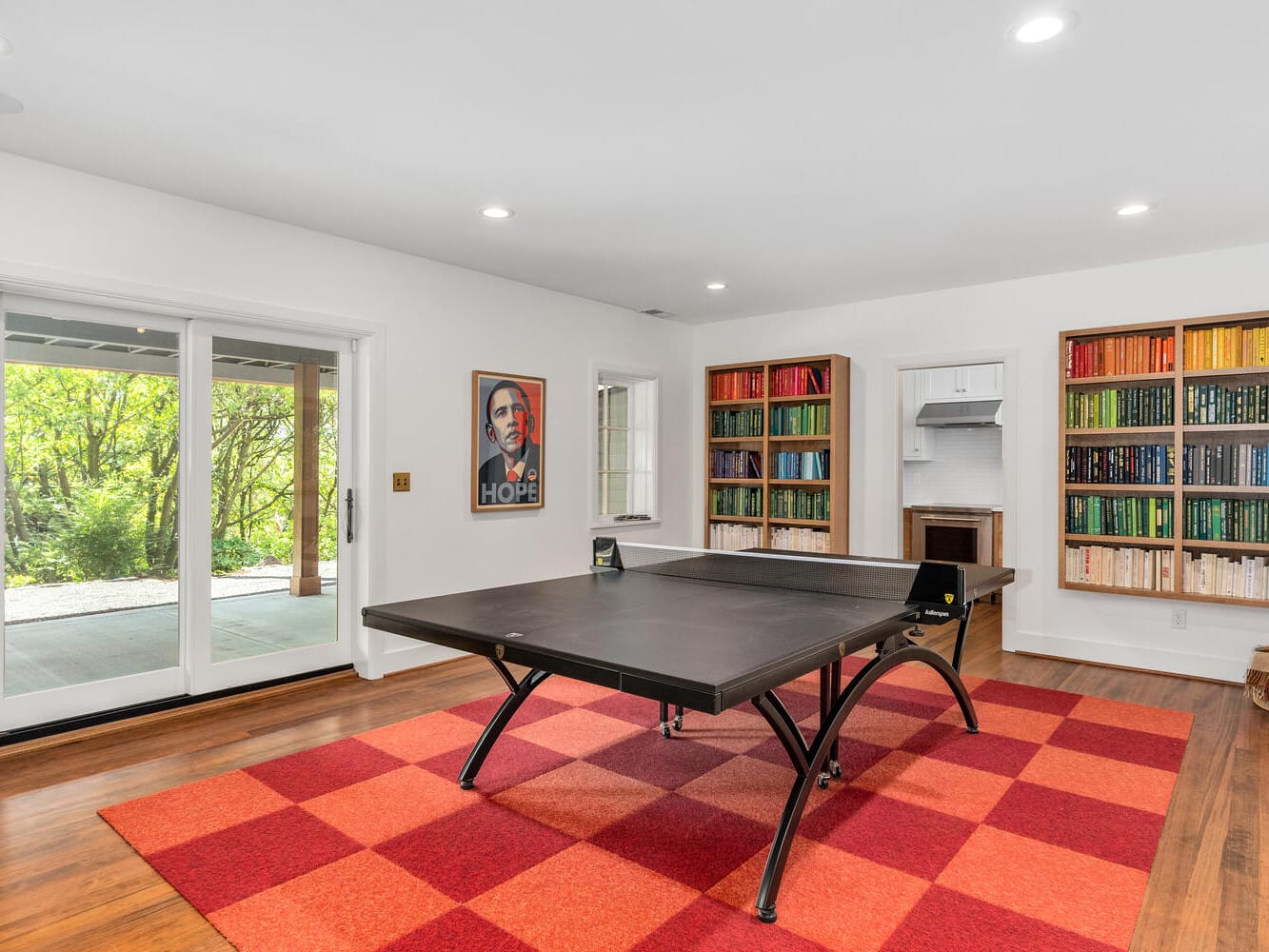 A modern room in Portland, Oregon boasts wooden flooring and a black ping pong table on a red checkered rug. Two colorful bookcases line the walls, while a large sliding door offers a garden view. A framed poster adorns the wall, adding to the stylish atmosphere.