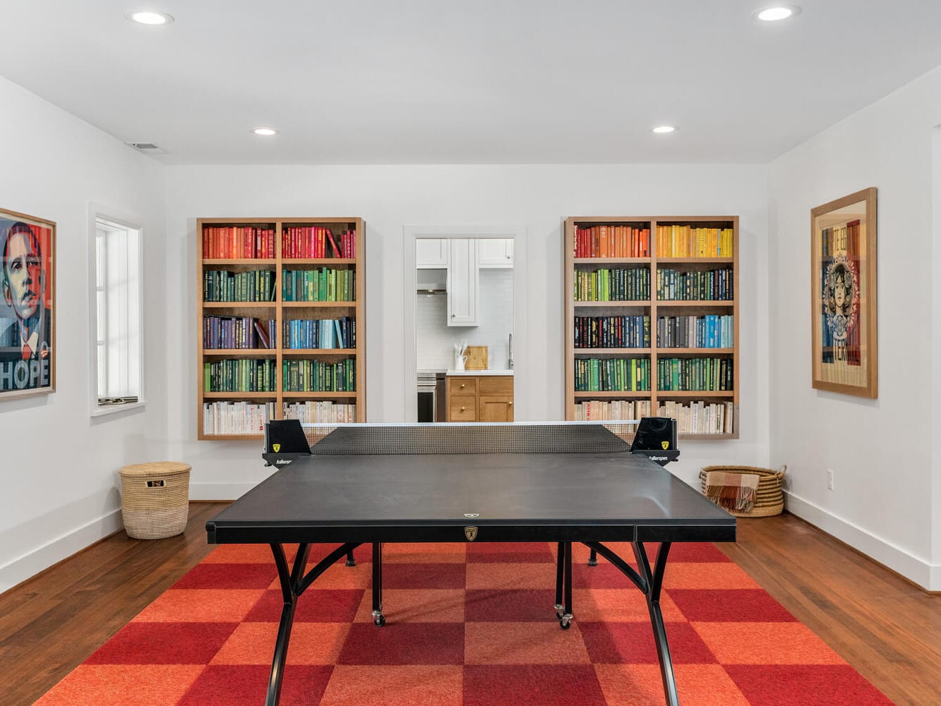 A room in Portland, Oregon features a table tennis setup on a red checkered rug. Colorful bookshelves and framed art adorn the white walls, while a small counter area is visible through a doorway, adding to the inviting charm of this vibrant space.