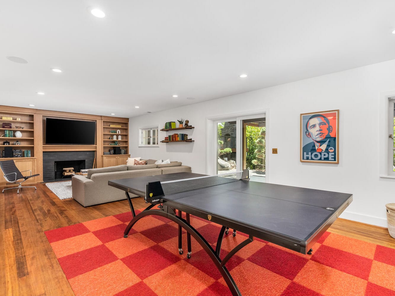 Spacious recreation room in Portland, Oregon, featuring a ping pong table on a red checkered rug. A beige sectional sofa faces a large TV above the fireplace, with built-in wooden shelves adorning the wall. A Hope poster is displayed, and large windows offer beautiful garden views.