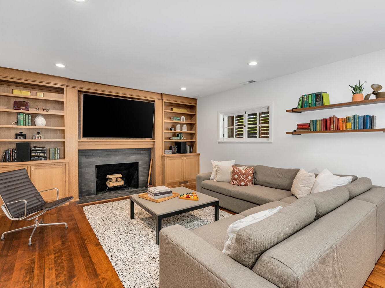 A cozy Portland, Oregon living room features a large gray sectional sofa, a modern black chair, and a wooden coffee table. Built-in bookshelves flanking the fireplace are adorned with books and plants. The walls are white, complementing the hardwood floor.