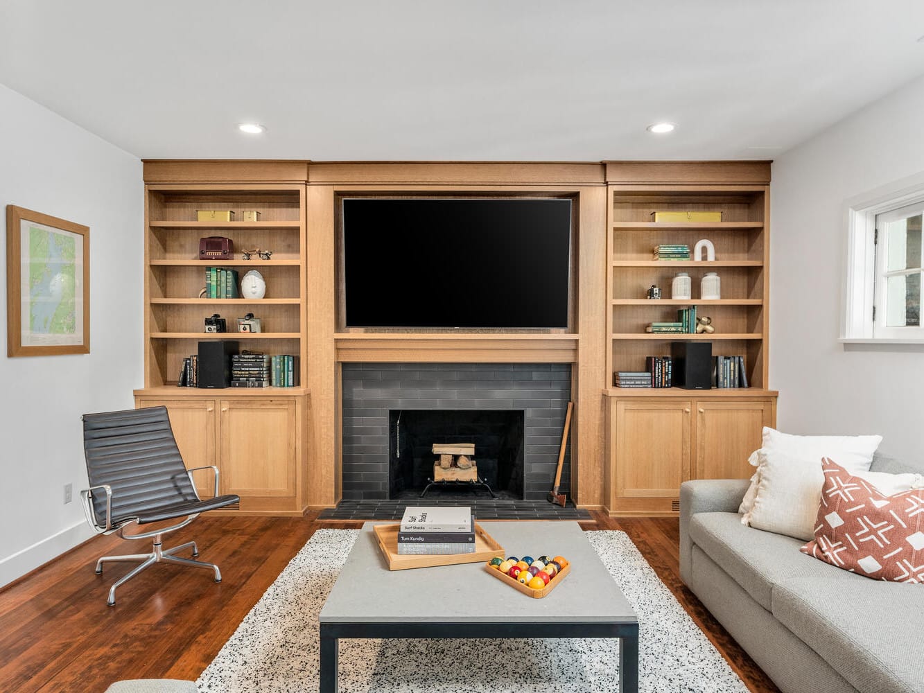 A modern living room in Portland, Oregon, features a large TV above a dark fireplace, flanked by wooden shelves filled with books and decor. A gray sofa and a sleek chair sit on a patterned rug around a low table. A small window and framed artwork adorn the walls.