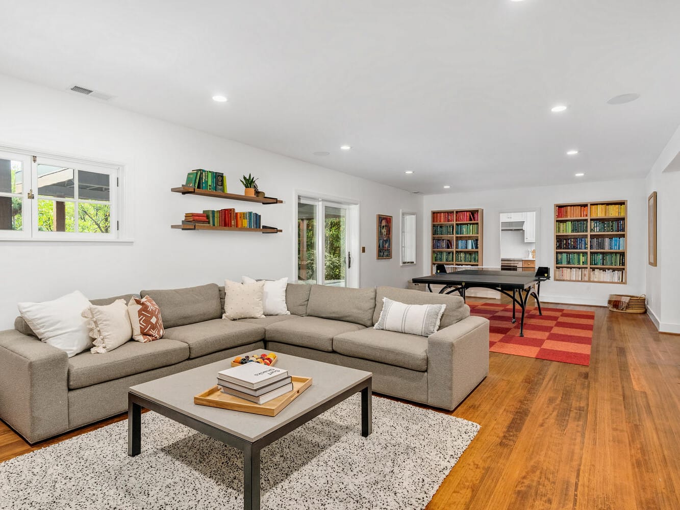 A spacious living room in Portland, Oregon, features a large beige sectional sofa, a coffee table with books and plants, wall-mounted shelves, and a view of a game area with a table tennis setup. The room has wooden floors and a colorful bookshelf.