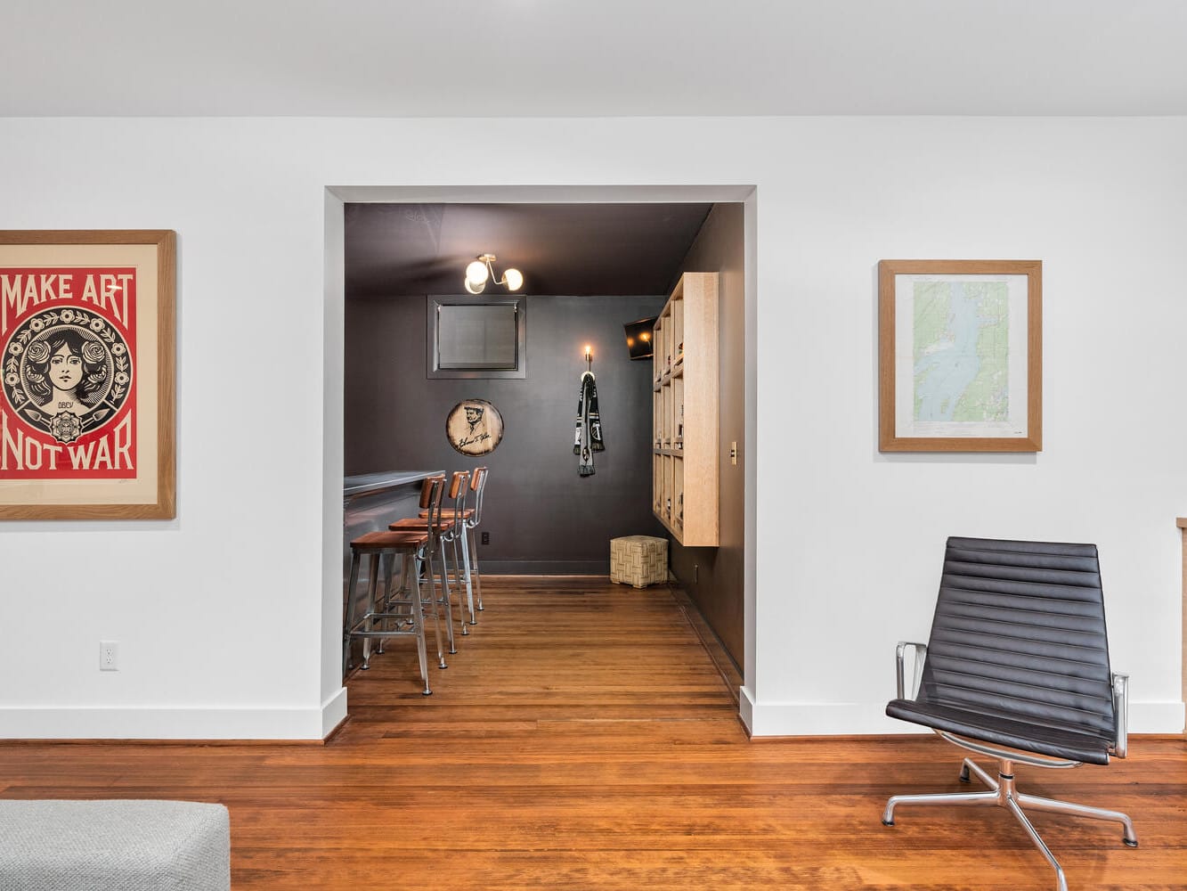 A room with wooden floors features a modern black chair and wall art, including a Make Art Not War poster. There's a bar nook with tall chairs and a framed Portland, Oregon map on the right wall. Shelves are partially visible on the far right.