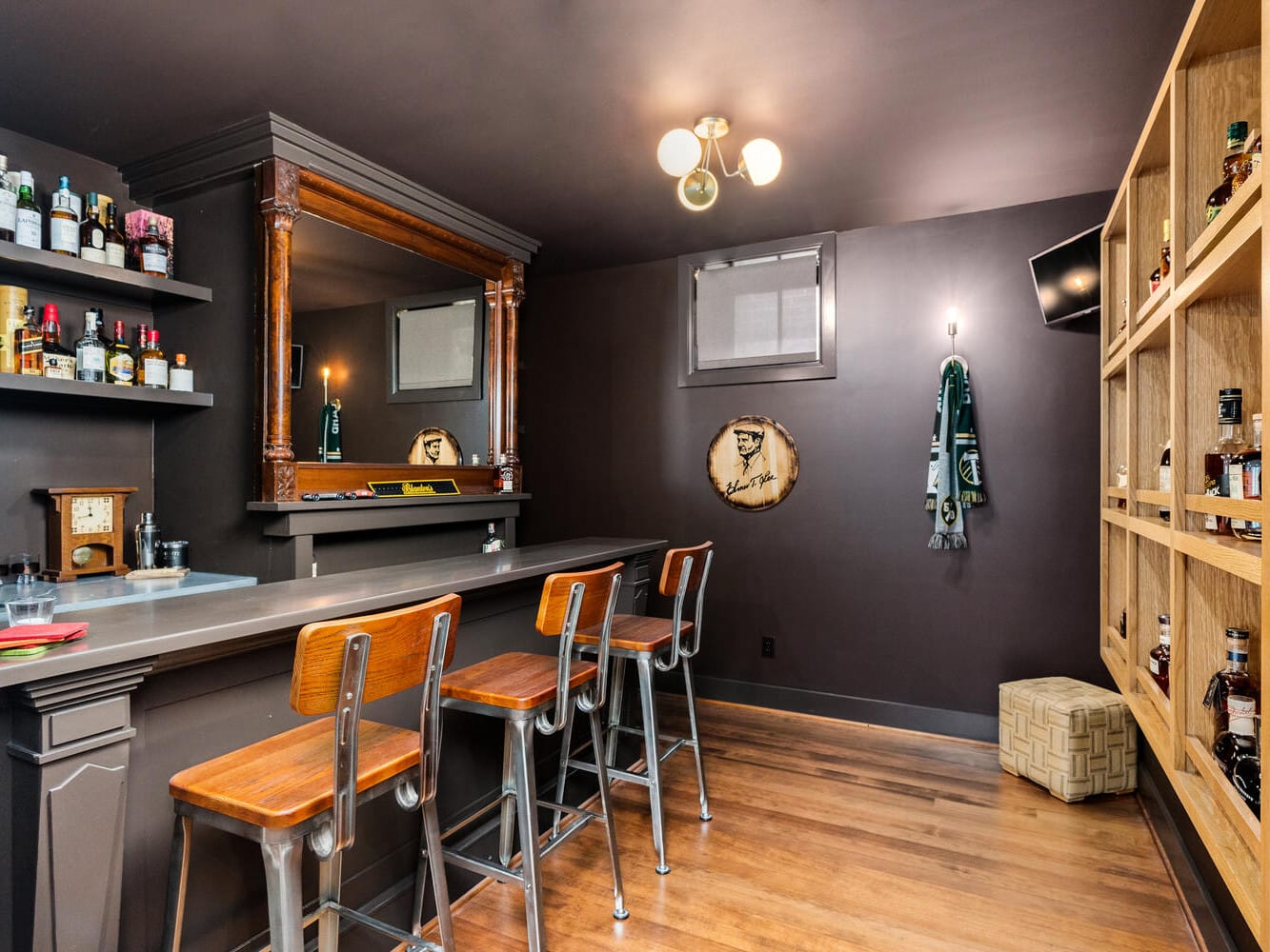 A cozy Portland, Oregon home bar with wooden stools and a gray counter. Bottles line the shelves and a mirror hangs on the wall. Warm lighting and a team logo scarf add to the ambiance. Wooden floors and shelving create a welcoming vibe.