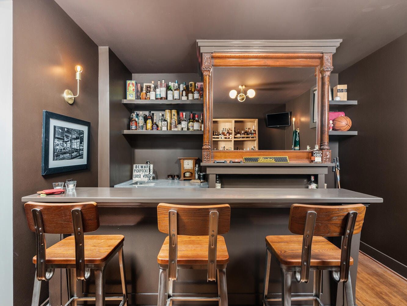 A cozy home bar in Portland, Oregon, features three wooden stools against dark walls and a wooden mirror panel. Shelves boast various bottles, while a small framed picture and basketball add unique decor. Soft yellow lighting casts a warm glow throughout the inviting space.