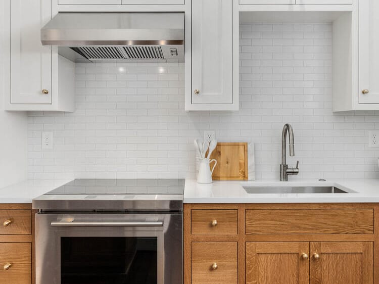 A modern kitchen in Portland, Oregon, featuring white upper cabinets, wooden lower cabinets, and a stainless steel oven with a built-in range hood. There's a white subway tile backsplash, a small sink with a faucet, and a cutting board with utensils nearby.