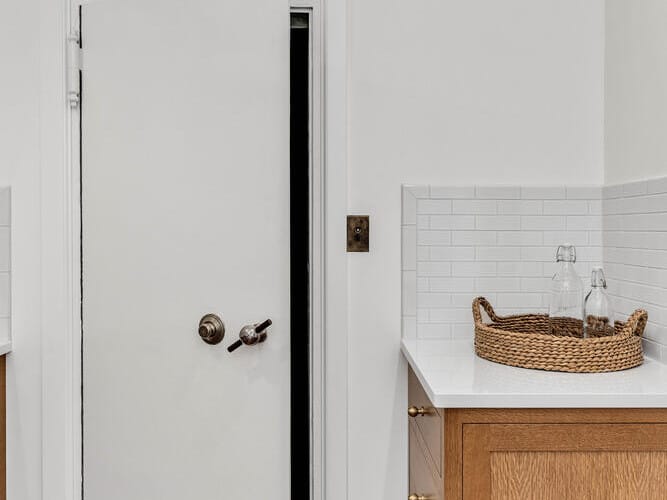 A modern kitchen in Portland, Oregon, features wooden cabinets with white countertops. Two upside-down glasses rest in a wicker tray. A door, slightly ajar, sits between the cabinets, revealing a dark interior. The walls are white with a tiled backsplash.