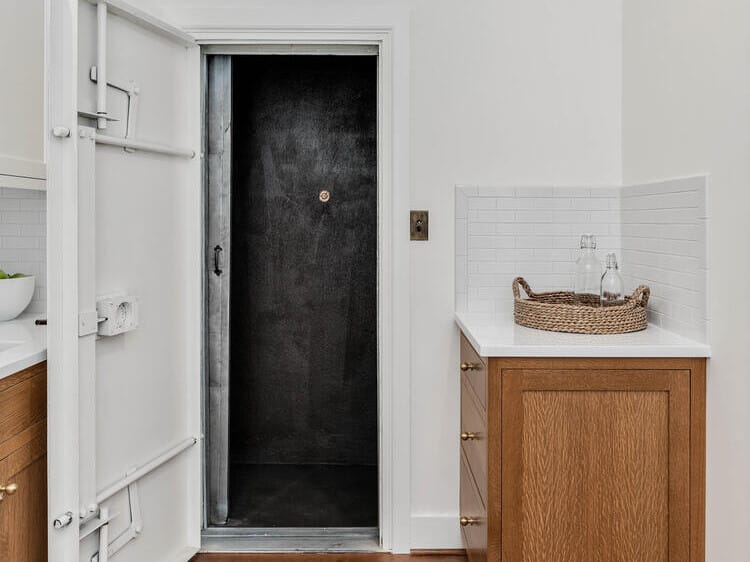 A room with wooden floors captures the charm of Portland, Oregon, featuring a small open vault with a heavy door. To the right, a wooden cabinet topped with a wicker basket and glassware sits gracefully against a white tiled backsplash.