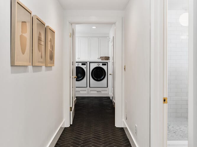 A narrow hallway with black herringbone tile flooring evokes a classic Portland, Oregon vibe as it leads to a laundry room. Inside, you'll find a washer and dryer, while beige framed abstract art adorns the left wall. A partially open door on the right reveals a tiled bathroom.