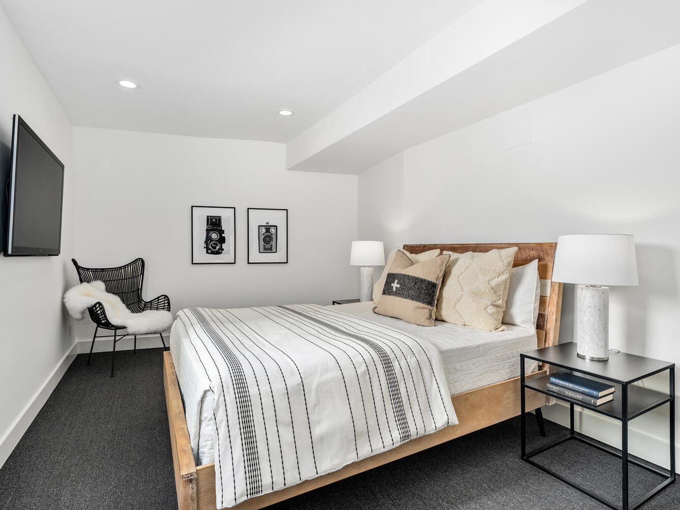 A modern bedroom in Portland, Oregon, boasts a wooden bedframe adorned with neutral bedding and pillows. A black armchair draped in white fur sits in the corner. Two framed pictures hang above the chair, while a flat-screen TV is mounted on the adjacent wall.