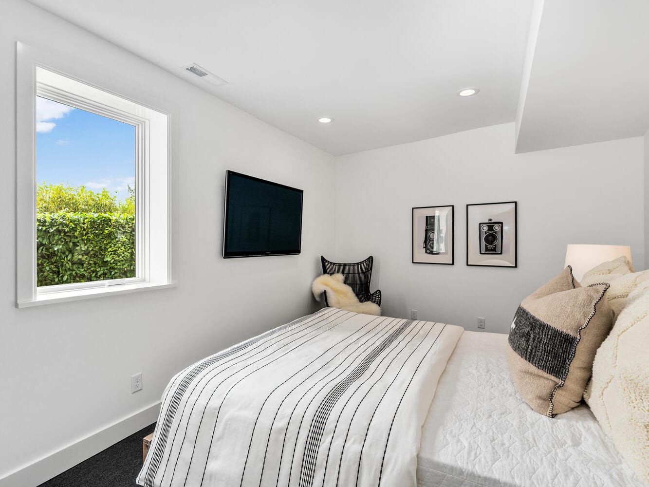 A modern bedroom in Portland, Oregon, features a neatly made bed with striped bedding and plush pillows. Opposite the bed is a wall-mounted TV. Framed pictures adorn the walls, while a window offers scenic views of greenery, allowing natural light to flood the space.