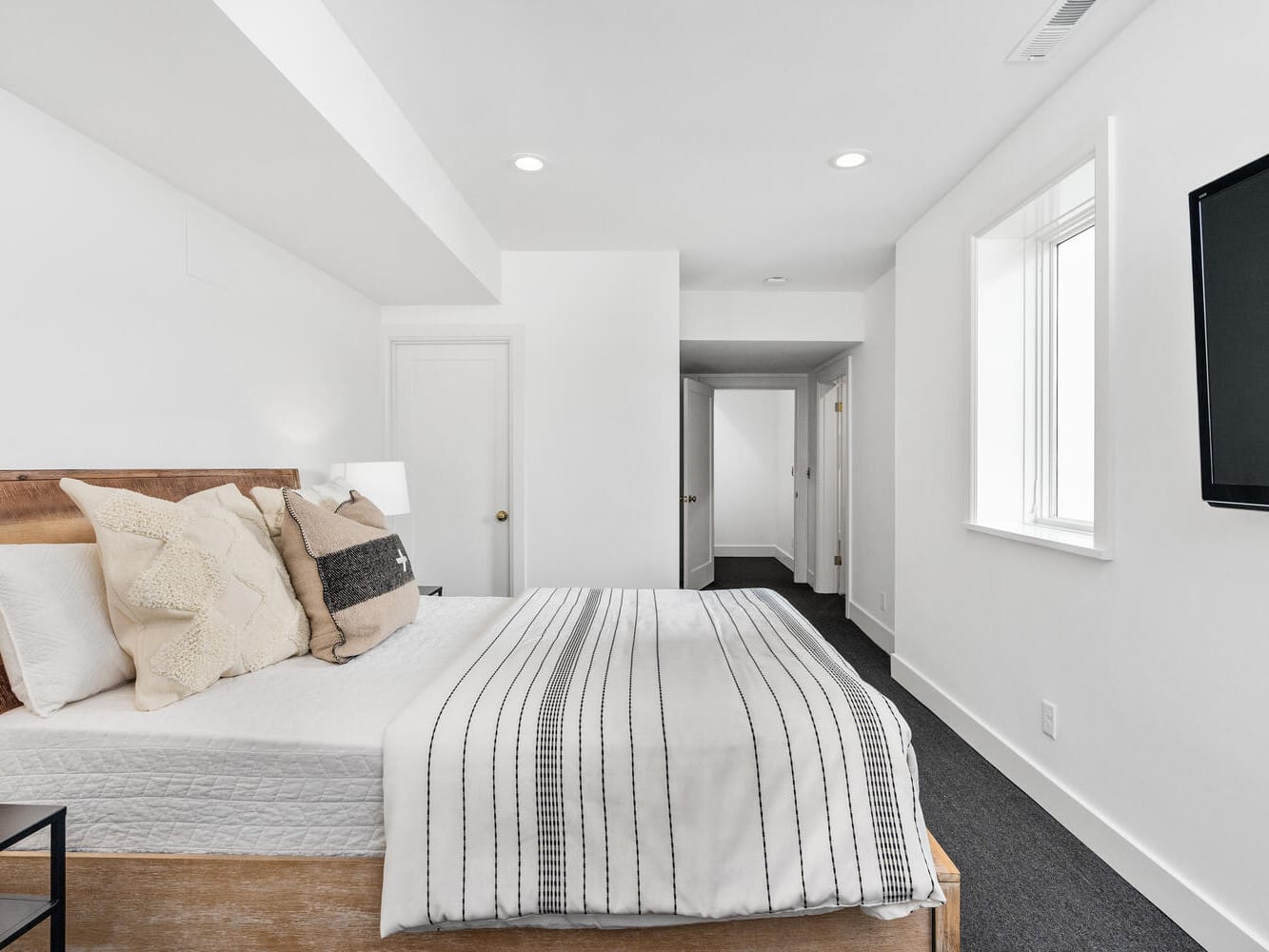 A minimalistic bedroom in Portland, Oregon, boasts a white bed adorned with striped bedding and a mix of white and beige pillows. The room features pristine white walls, a flat-screen TV mounted on the wall, and a dark carpet. Natural light pours in through a window on the right.