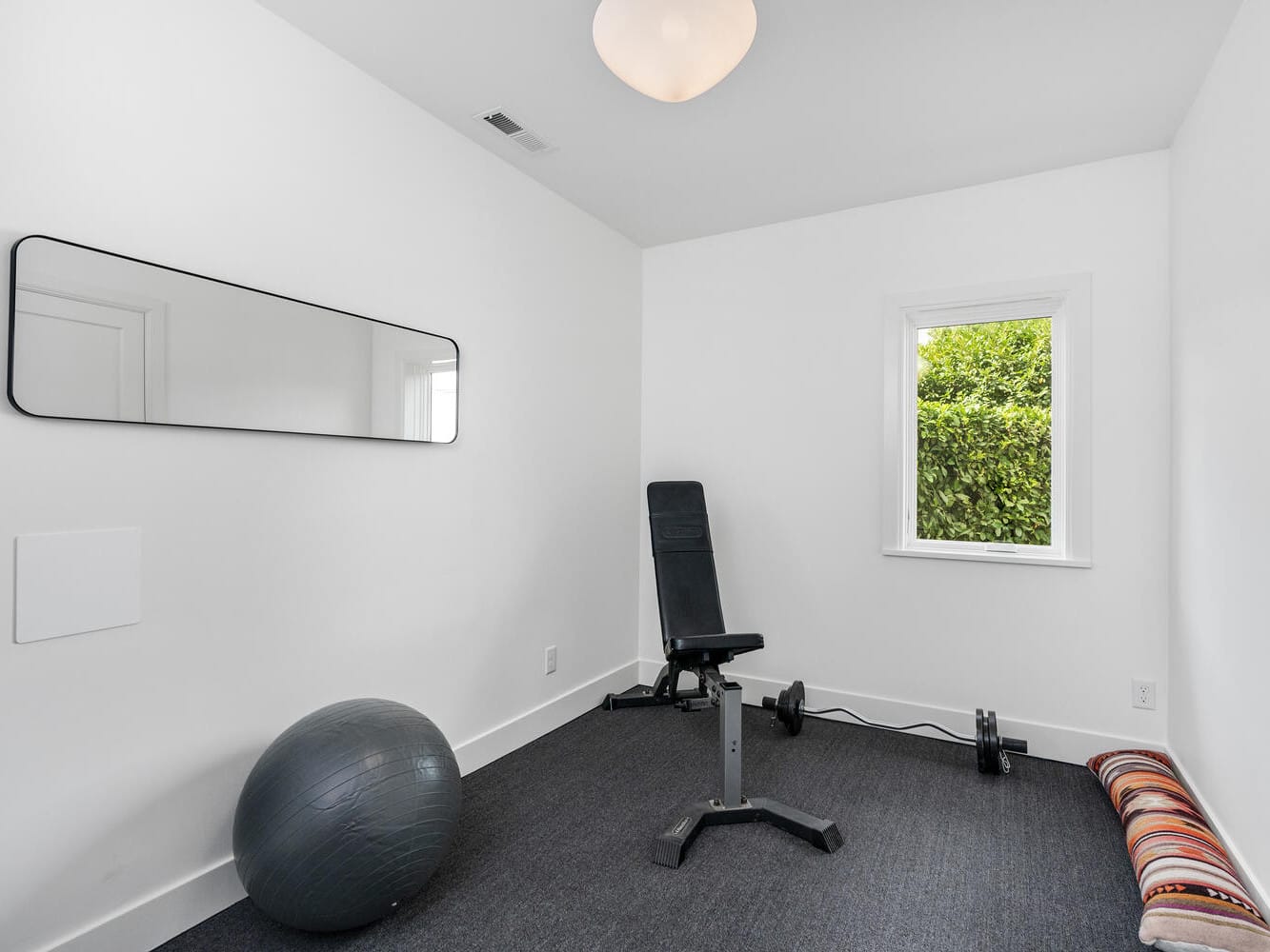 A minimalist home gym in Portland, Oregon features a black exercise bench, a stability ball, dumbbells, and a striped cushion on the floor. The room's white walls, rectangular mirror, single window, and gray carpeting establish a serene setting under the round ceiling light.