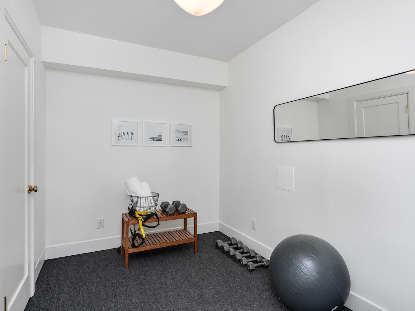 A minimalist home gym in Portland, Oregon features a wooden bench holding towels and dumbbells. A gray exercise ball and more dumbbells rest on the carpeted floor. Three framed photos adorn the wall, complemented by a large horizontal mirror.