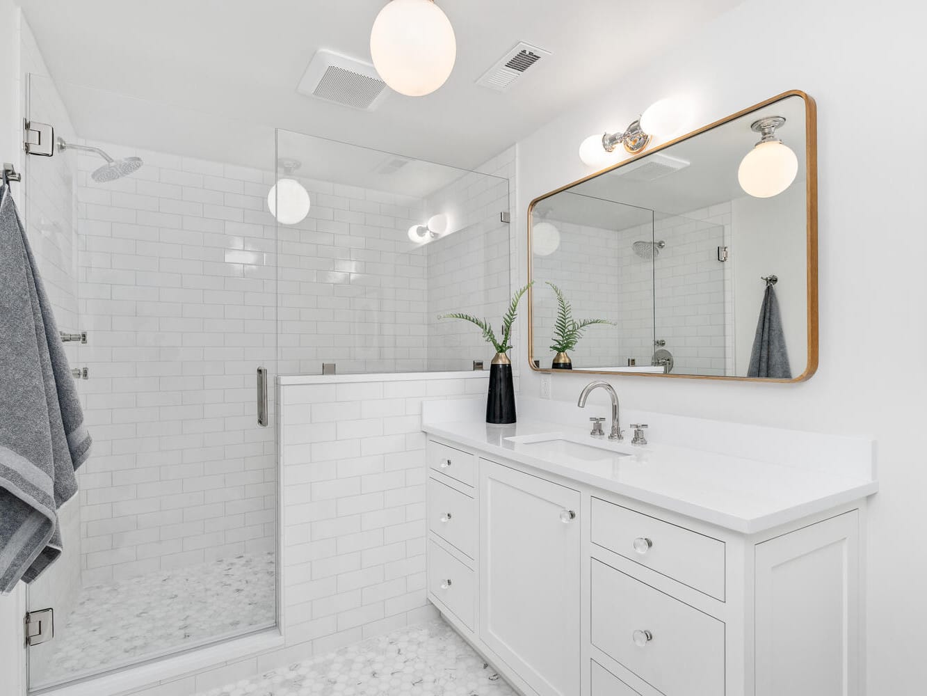 A modern bathroom in Portland, Oregon, showcases white cabinets and a large mirror above the sink. On the left is a walk-in glass shower with white subway tiles. The floor features a hexagonal pattern, while a black vase with green leaves adds elegance to the counter.