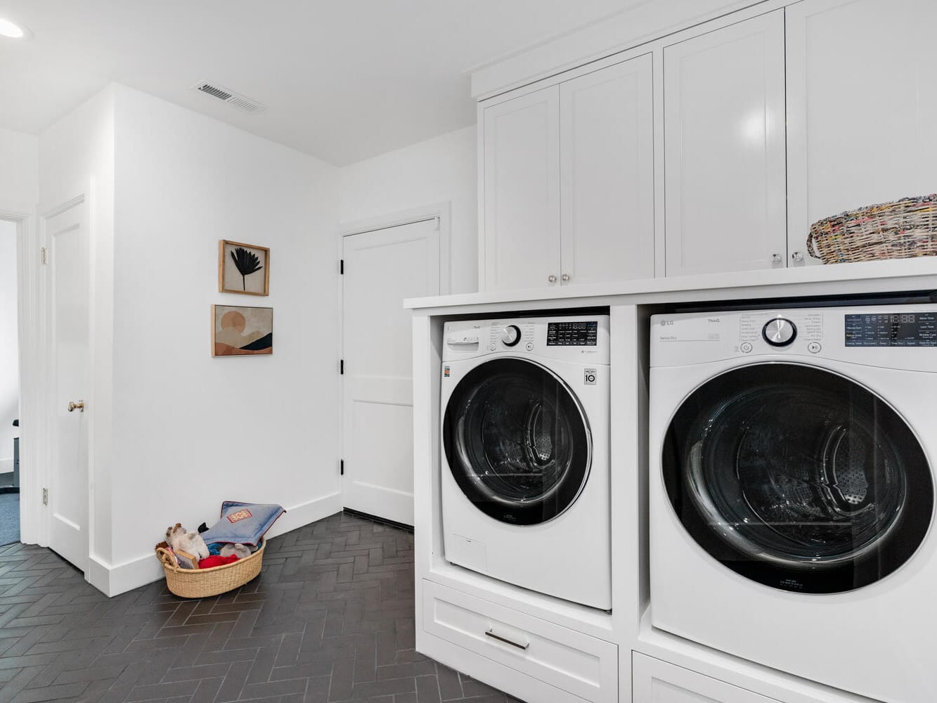 A modern laundry room in Portland, Oregon, features a front-loading washer and dryer. Above the appliances are sleek white cabinets, while a woven basket sits on the counter. The room boasts herringbone-patterned dark flooring and white walls, with two wall artworks and a small pet basket nestled in the corner.