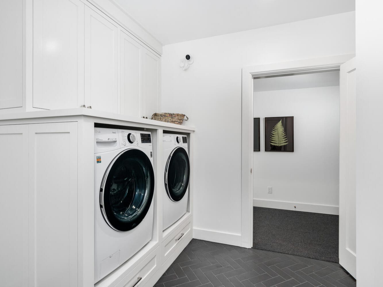 A modern laundry room in Portland, Oregon, boasts white cabinetry with a front-loading washer and dryer. A basket rests on the countertop, while dark herringbone-patterned tiles line the floor. An open door leads to a hallway adorned with framed artwork.