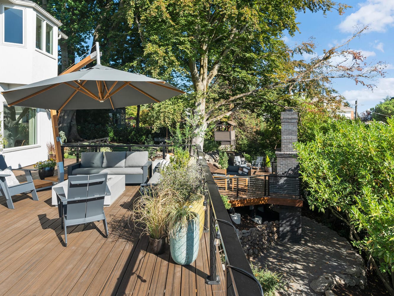 A spacious wooden deck with modern gray outdoor furniture, shaded by a large umbrella and reminiscent of Portland, Oregon's natural charm, features potted plants and a cozy fire pit surrounded by trees. The space offers serene views of lush greenery under a clear blue sky.