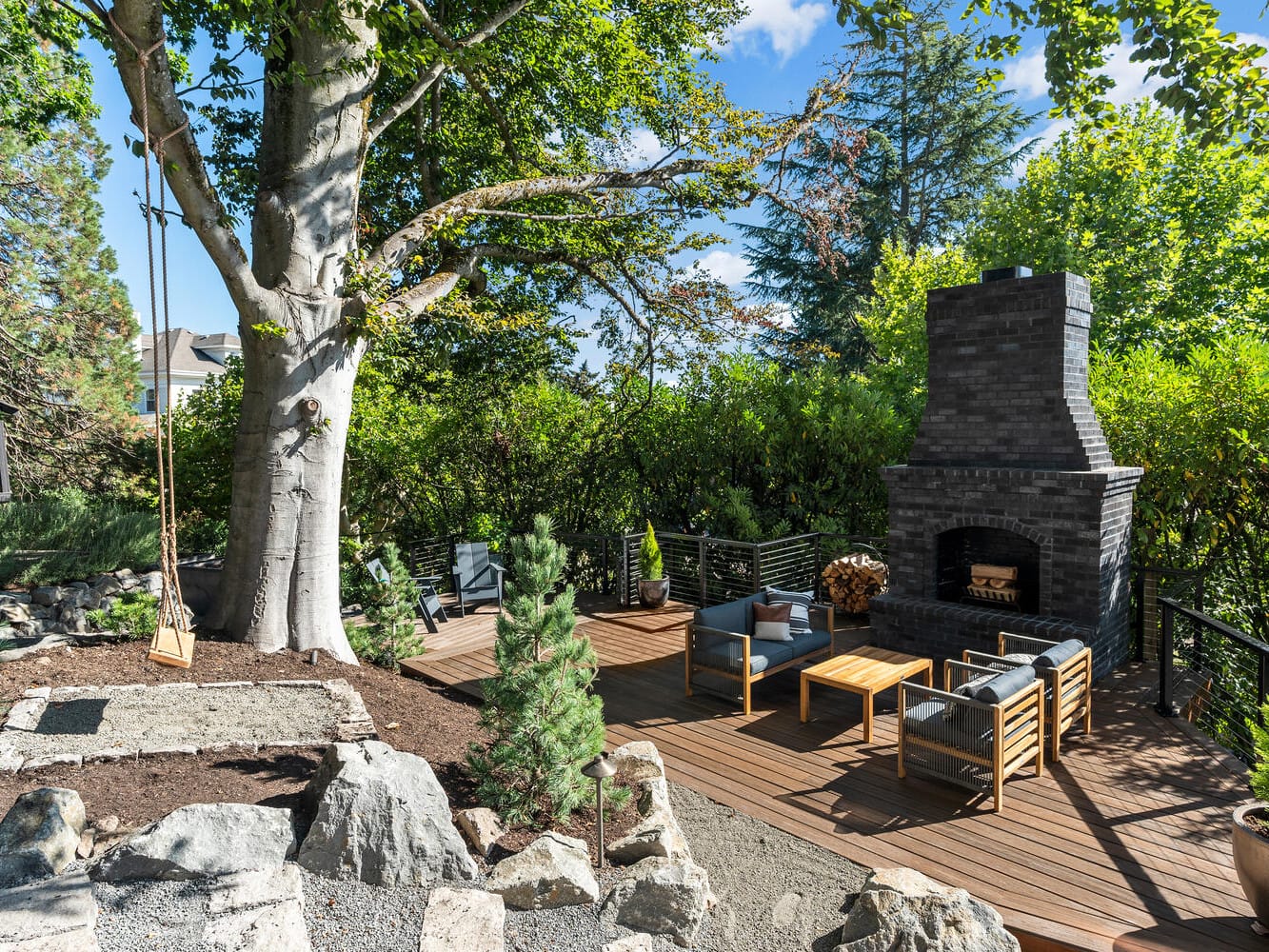 An outdoor patio in Portland, Oregon, boasts a wooden deck with black metal railing. It features a stone fireplace, seating with gray cushions, and a wooden swing hanging from a large tree, surrounded by lush greenery and rocks. It's a bright and sunny day.