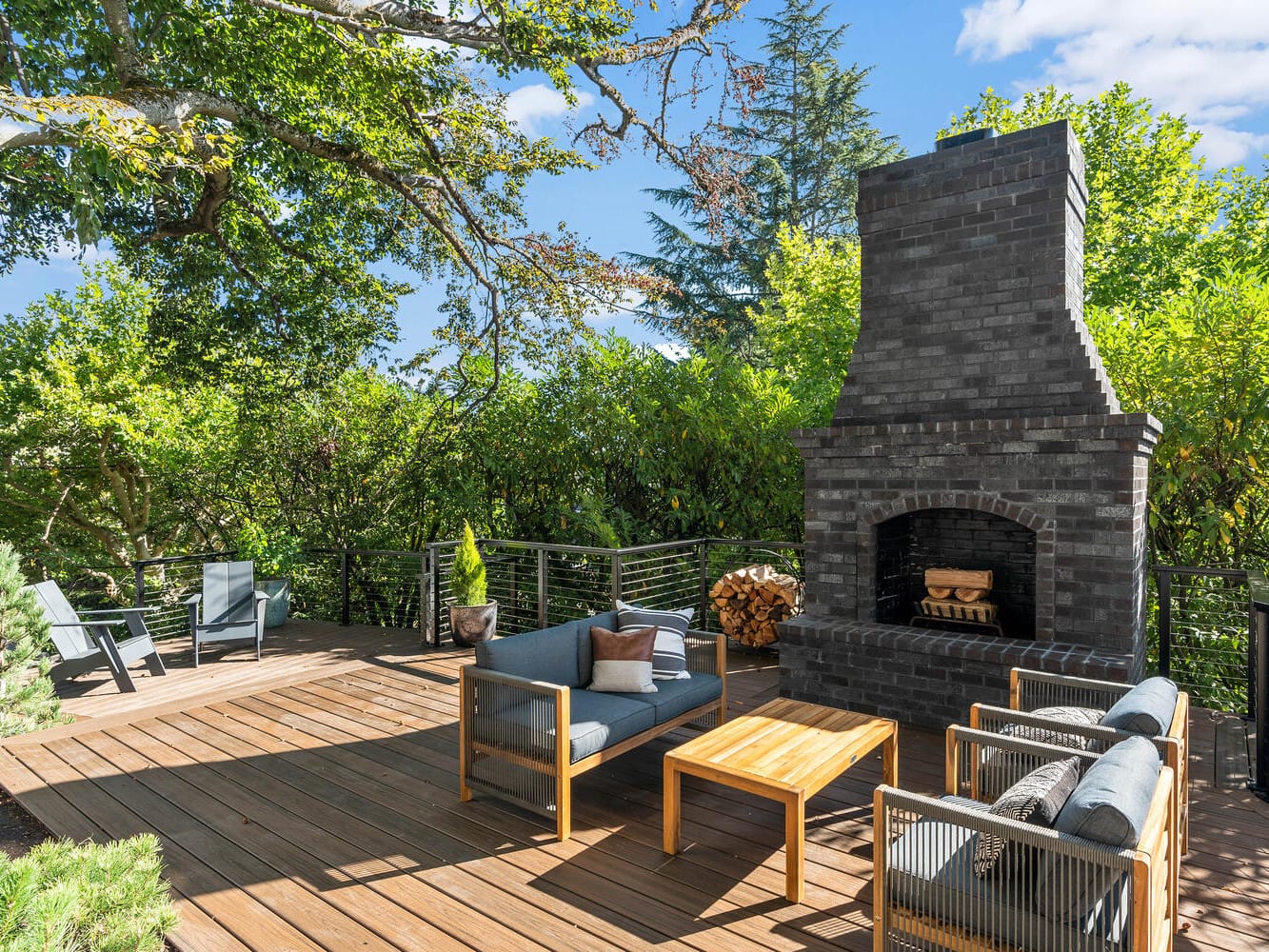 Wooden deck with outdoor seating, a brick fireplace, and surrounding lush greenery reminiscent of Portland, Oregon. Blue sky with scattered clouds above creates a cozy, inviting setting for relaxation or gatherings.