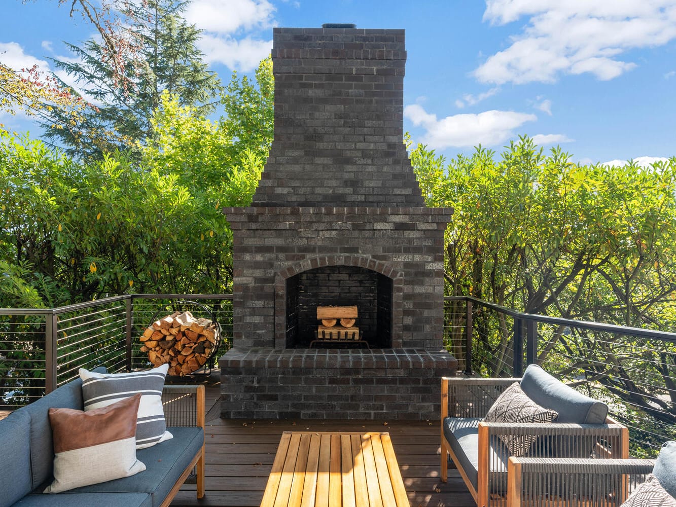 A cozy outdoor patio in Portland, Oregon features a dark brick fireplace surrounded by lush greenery. Two cushioned seats with pillows flank a wooden table, offering a perfect spot to unwind. Nearby, a stack of firewood hints at cozy evenings under the blue sky dotted with scattered clouds.