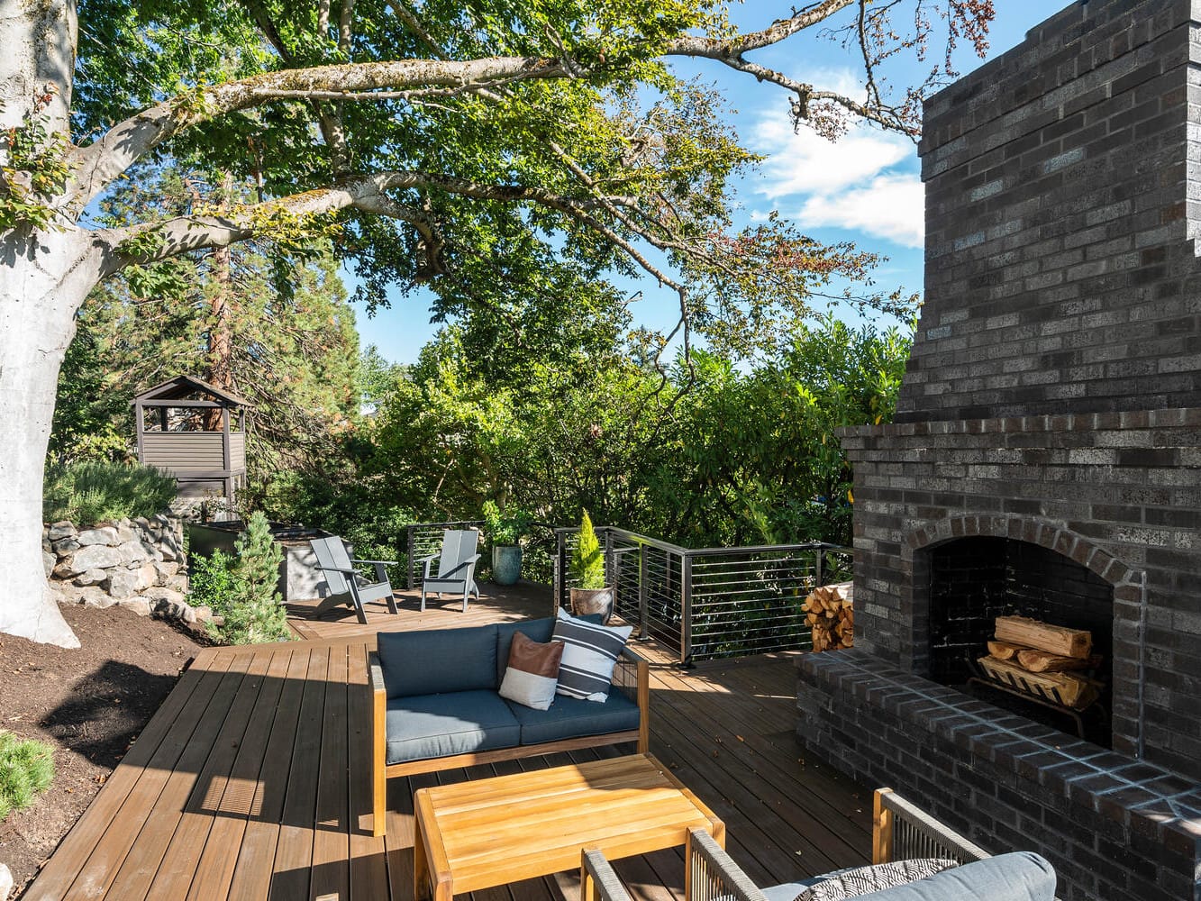 A wooden outdoor deck in Portland, Oregon, features a large black brick fireplace surrounded by trees. Cushioned benches and a wooden table offer seating, while lounge chairs are seen in the background next to a wooden playhouse under the clear blue sky.