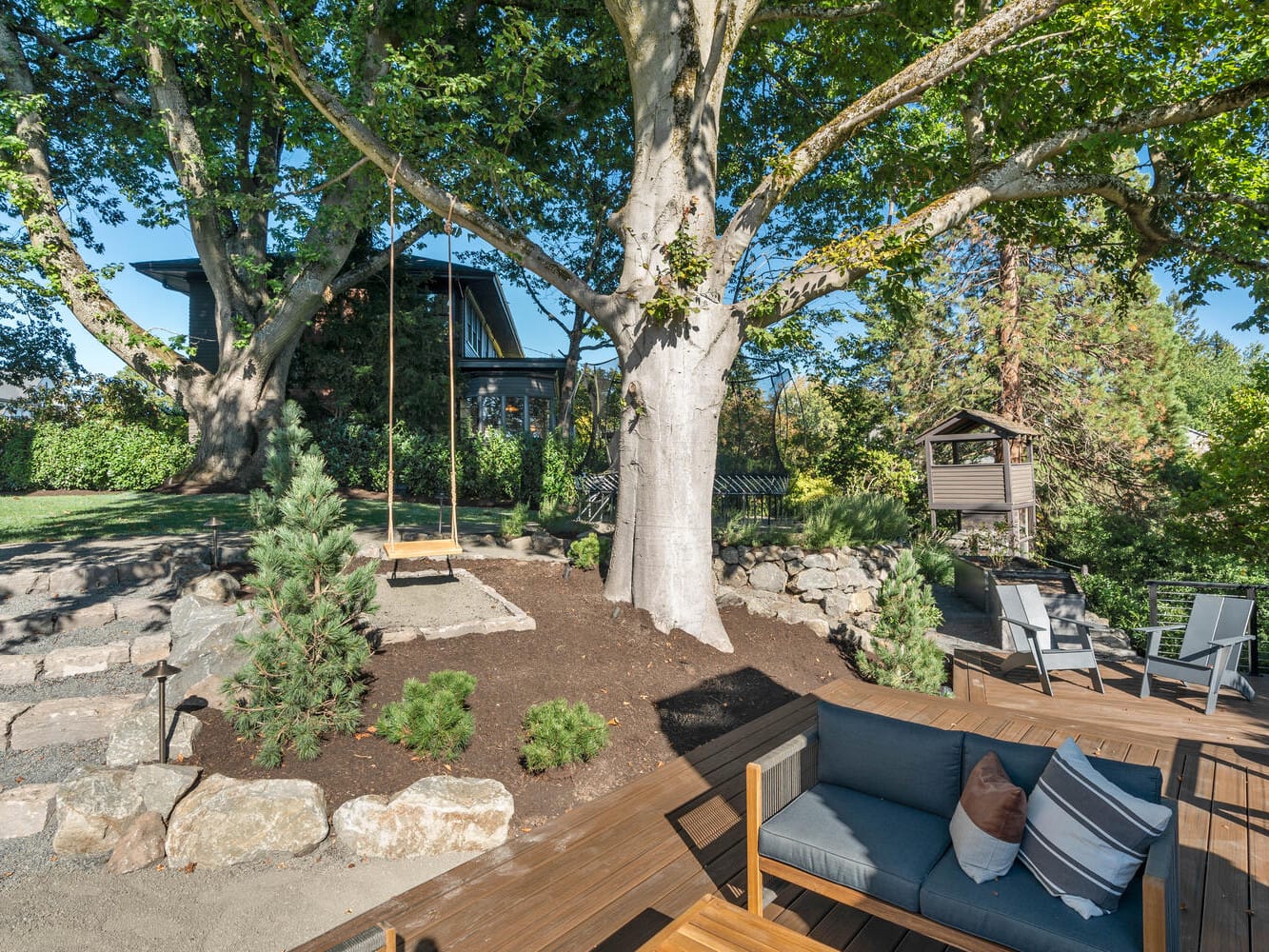 In a charming Portland, Oregon backyard, a large tree hosts a delightful swing. A wooden deck boasts outdoor furniture, including a gray sofa and tables. The surroundings are lush with greenery, and in the background stands a small structure, possibly a cozy playhouse.