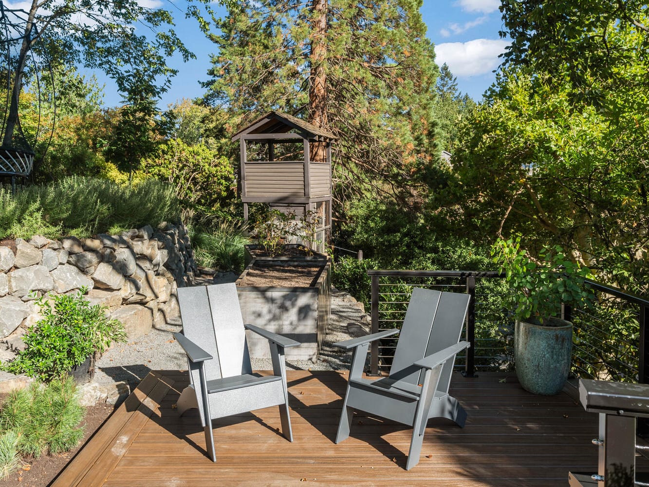 A wooden deck with two gray chairs overlooks a lush garden in Portland, Oregon. A small, elevated treehouse is in the background, surrounded by tall trees and rocks. A large potted plant adds to the greenery on the deck. The scene is sunlit and serene.