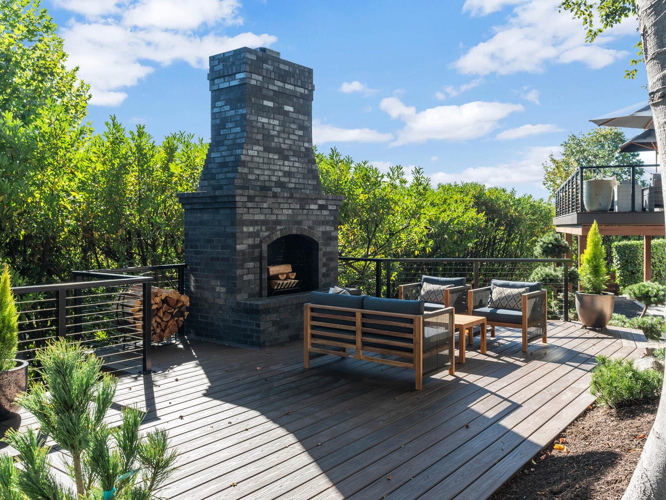 A spacious outdoor deck with a tall, dark brick fireplace evokes the charm of Portland, Oregon. Two wooden sofas with dark cushions face each other around the fireplace. Lush greenery surrounds the deck under a clear blue sky.