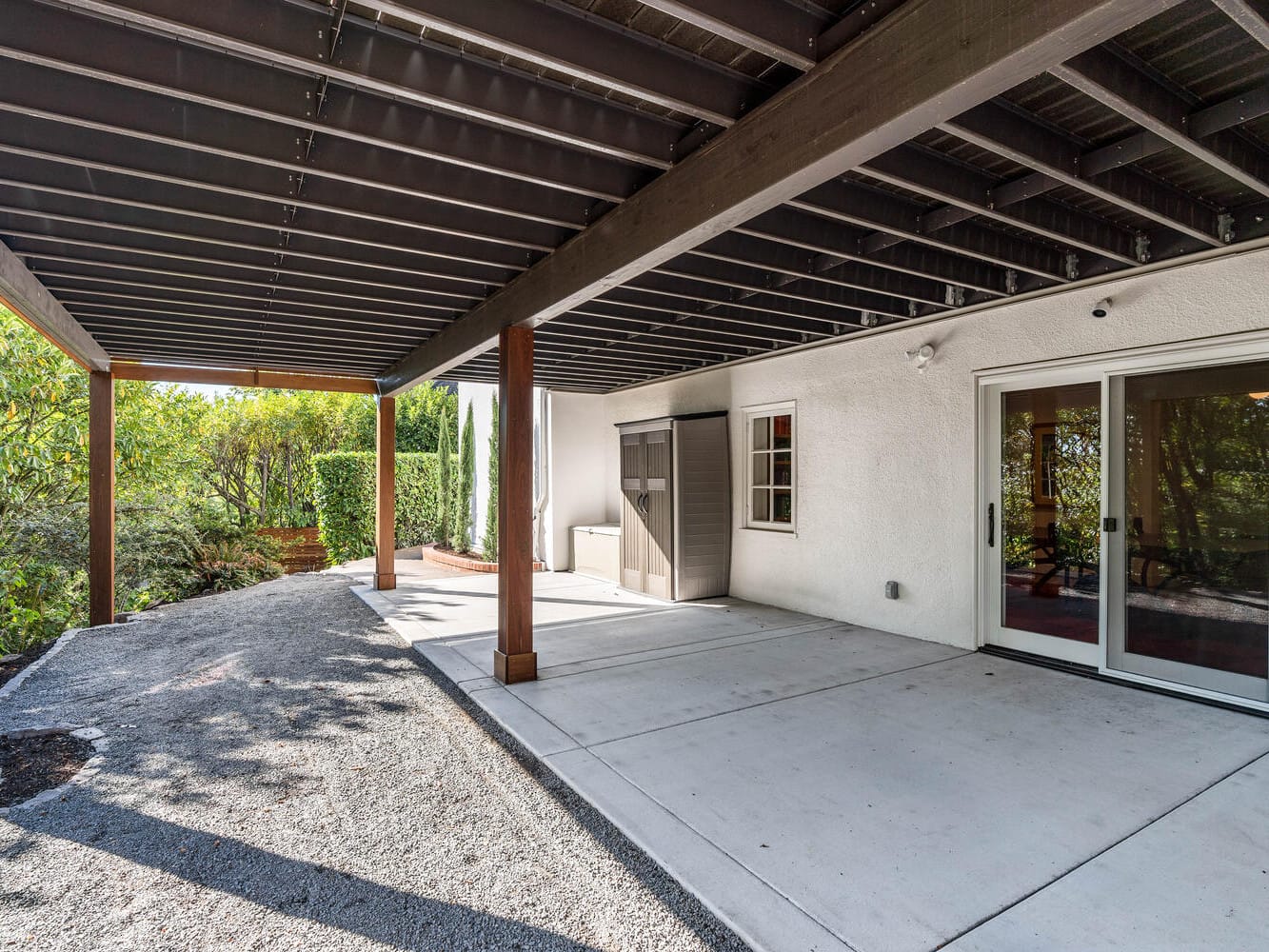 A covered patio with a concrete floor and wooden supports extends from a white building in Portland, Oregon. Glass sliding doors lead inside, and a small window is visible. Bushes and greenery flourish along the edge of the patio, adding to its charm.