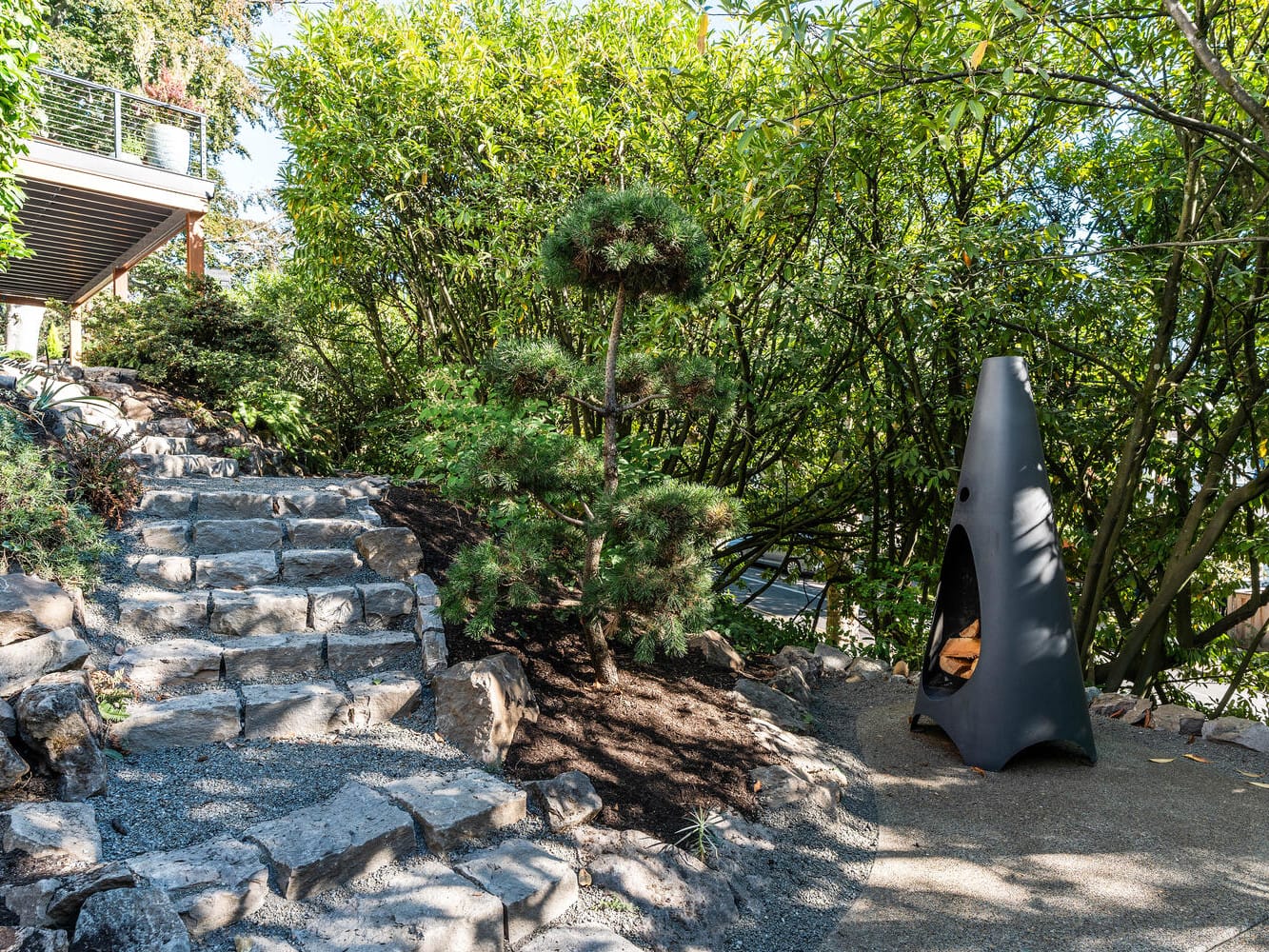 A serene garden scene in Portland, Oregon, features a stone pathway leading to a small pine tree surrounded by lush greenery. To the right, a modern black outdoor fireplace sits on a concrete slab, adding a contemporary touch to the natural setting.