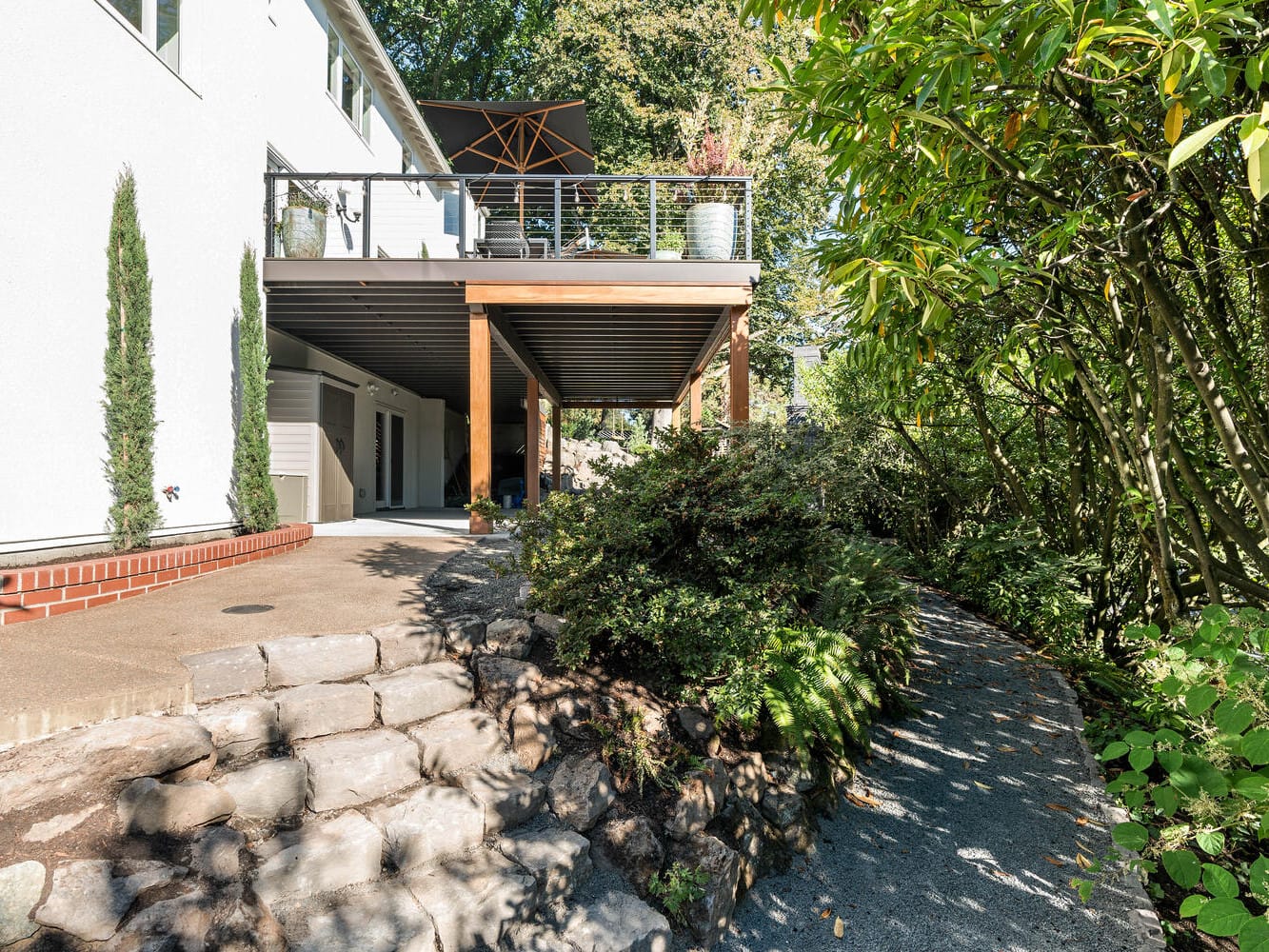 A two-story house in Portland, Oregon, with a white exterior boasts a wooden deck on the upper level, supported by pillars. A stone path and lush greenery surround the area, complemented by a sunshade and potted plant visible on the deck.