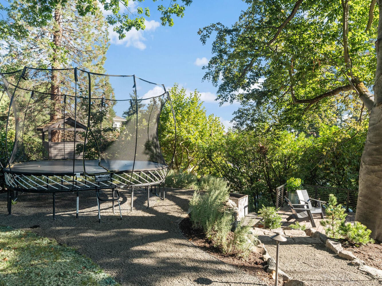 A backyard in Portland, Oregon, features a trampoline surrounded by trees and bushes. The ground is carpeted with small gravel stones, and a few wooden chairs rest on a stone patio to the right. Sunlight filters through the lush greenery, creating a serene atmosphere.