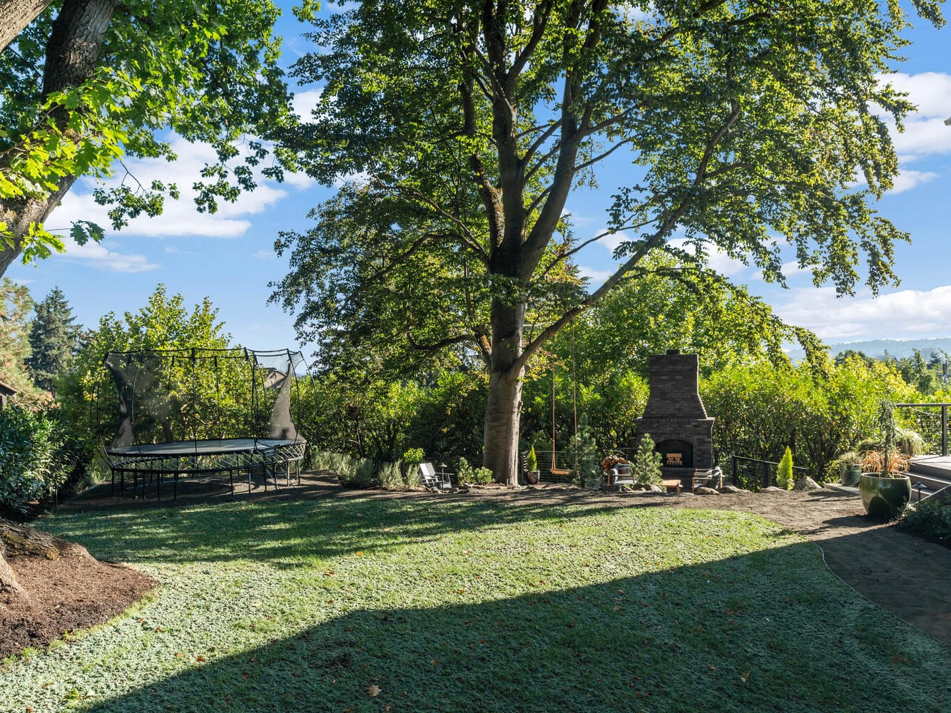 A lush Portland, Oregon backyard features a large tree providing shade. There's a trampoline to the left and an outdoor fireplace with seating to the right. The area is surrounded by dense greenery and a blue sky overhead.