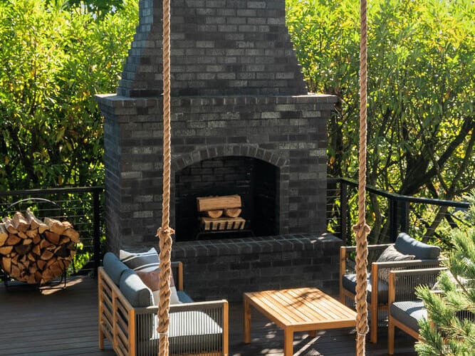 Outdoor patio in Portland, Oregon, featuring a tall dark brick fireplace, two cushioned chairs, and a wooden table on a wooden deck. A rope swing hangs in the foreground, surrounded by lush greenery under a blue sky.