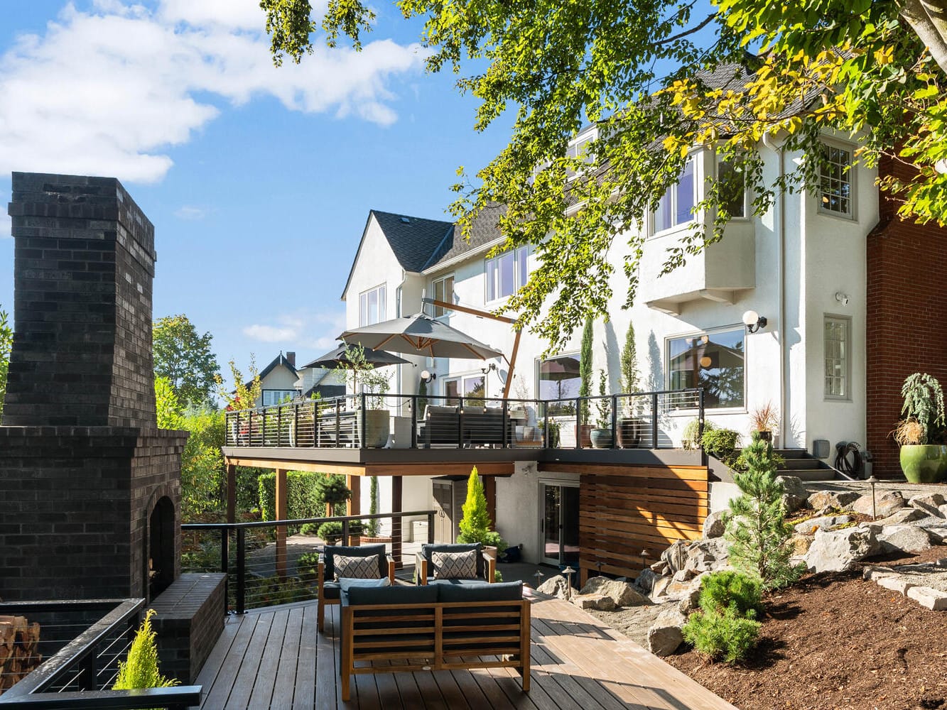 Backyard view of a modern two-story house in Portland, Oregon, with large windows and a spacious wooden deck. The deck features seating, a dining area under an umbrella, and is surrounded by trees and a brick outdoor fireplace. Sunny weather enhances the bright atmosphere.